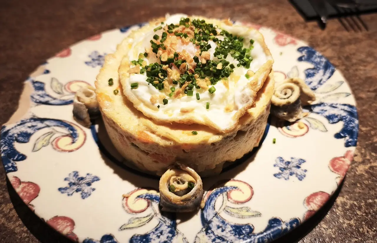 Layered dish topped with fried egg and chives, plated with rolled anchovies