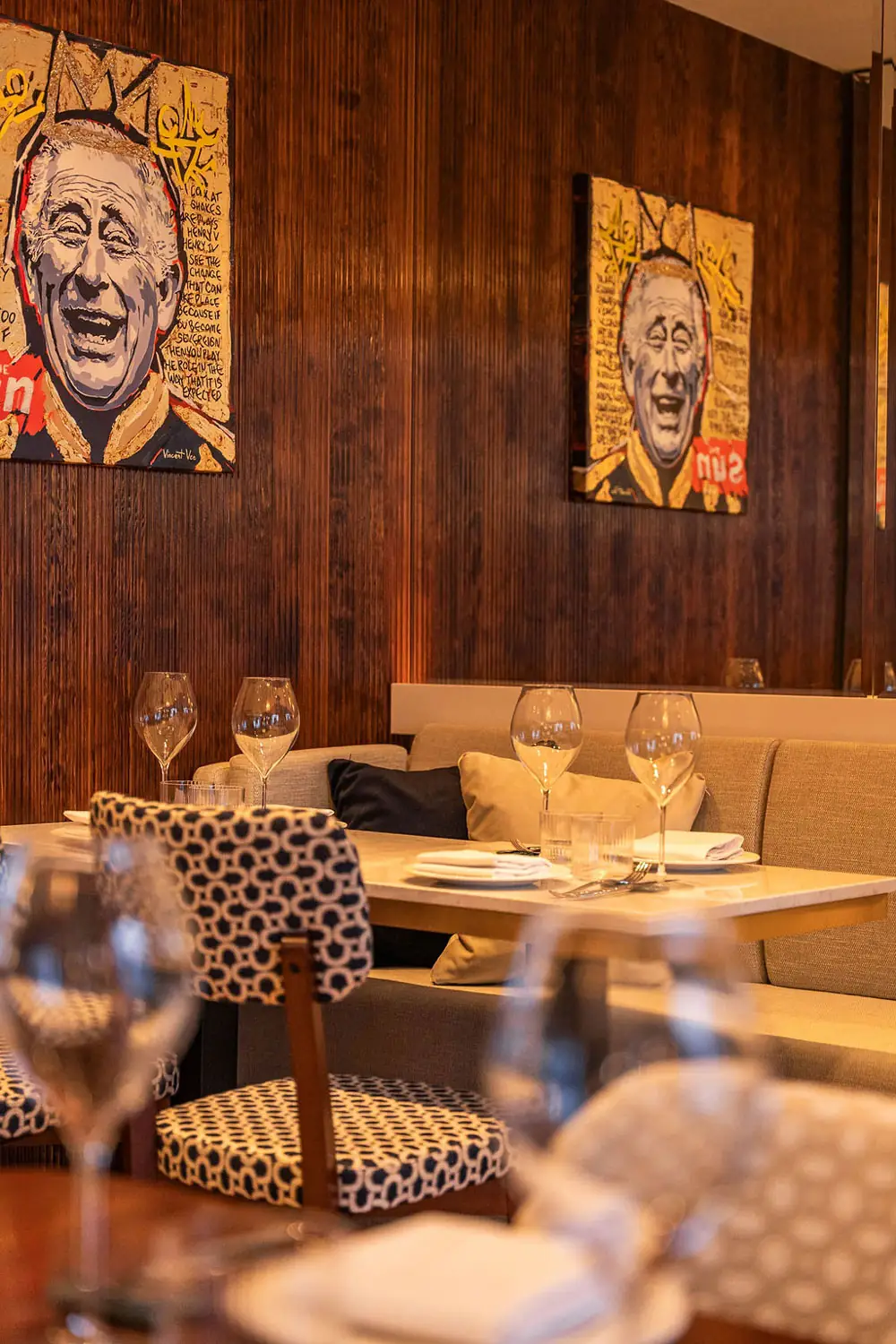 Restaurant dining nook with dark wood-panel walls hung with pop-art portrait paintings, a marble table set with wine glasses and plates and a patterned banquette