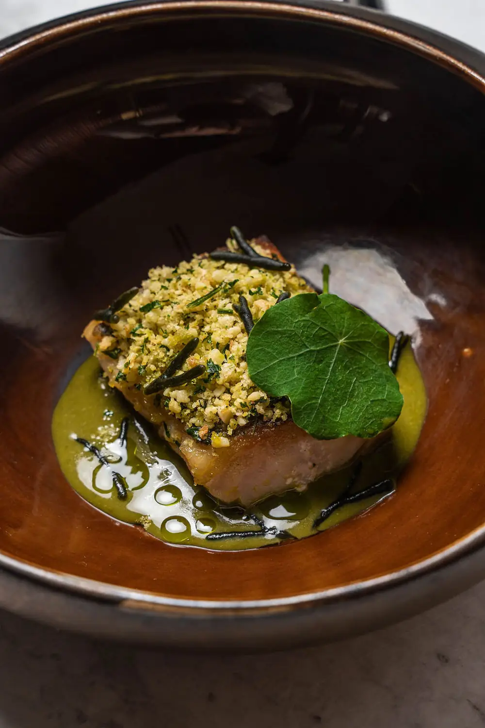 Herb-and-nut-crusted fish fillet on green sauce garnished with a nasturtium leaf, plated in a glossy brown ceramic bowl