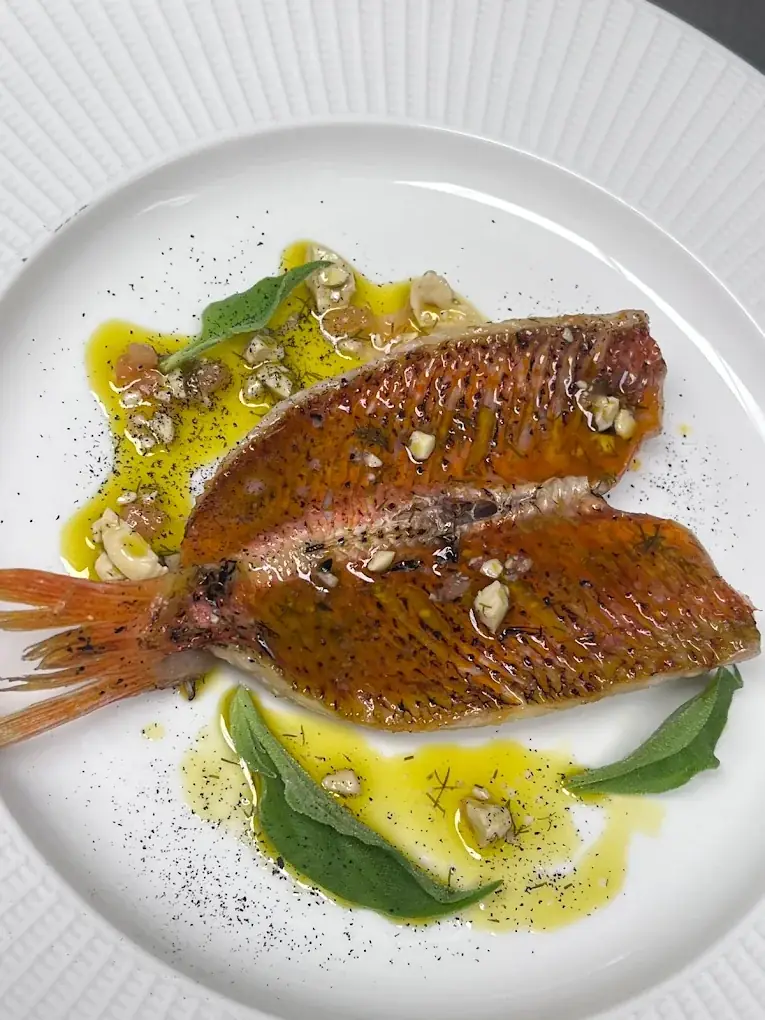 Whole grilled red mullet fish plated with charred lemon and herbs on a white plate.