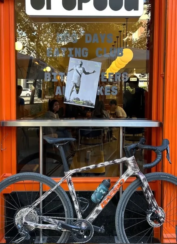 Road bike with a bidon parked against Grupeta's orange storefront window, diners at tables visible through the glass behind window lettering reading Dog Days Eating Club