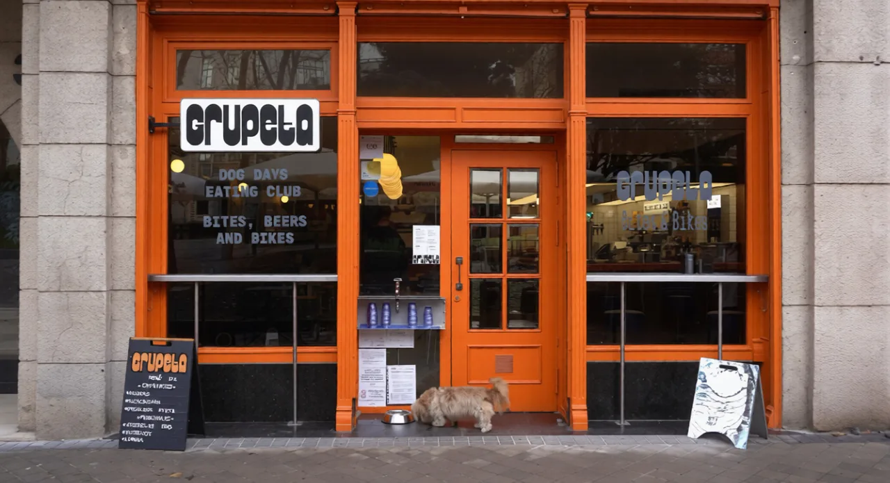 Full orange-framed facade of Grupeta with its black-and-white sign, chalkboard A-boards either side of the door and a small shaggy dog drinking from a metal bowl on the pavement