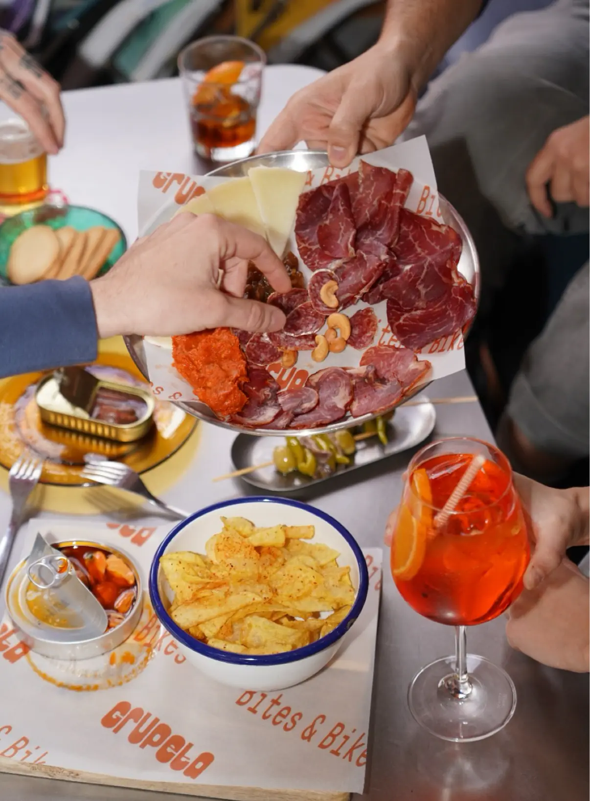 Table spread of cured meats, cheese and cashews, crisps, tinned seafood and a spritz cocktail