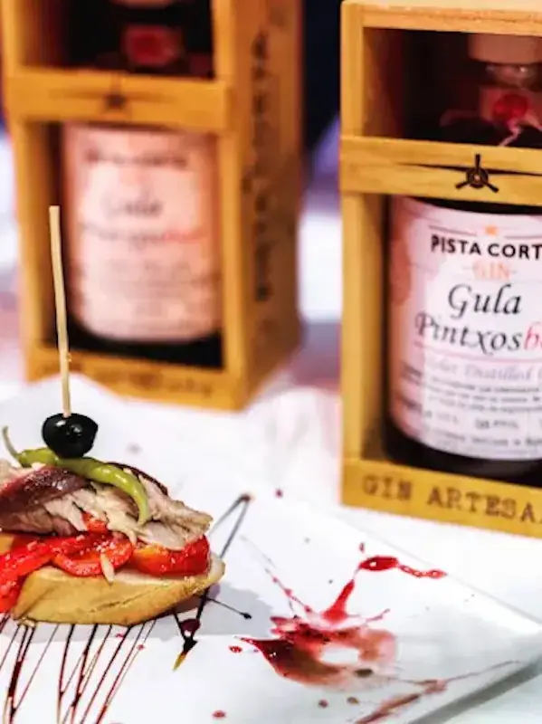 Skewered pintxo of cured meat, green guindilla pepper and a black olive on a slice of bread with red pepper, plated beside branded Gula Pintxosbar gin bottles in wooden gift cases.