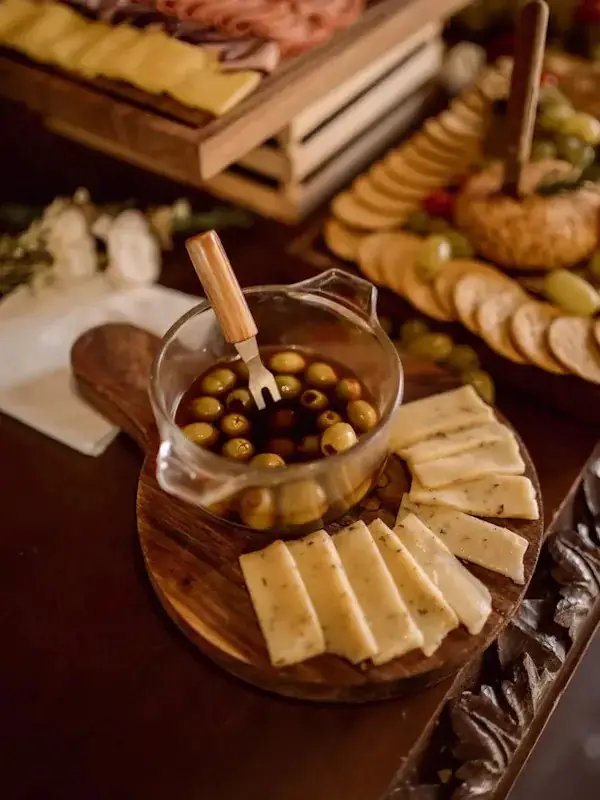 Wooden board with sliced cheese and a glass dish of marinated olives with a serving fork, crackers and cured meat visible behind.