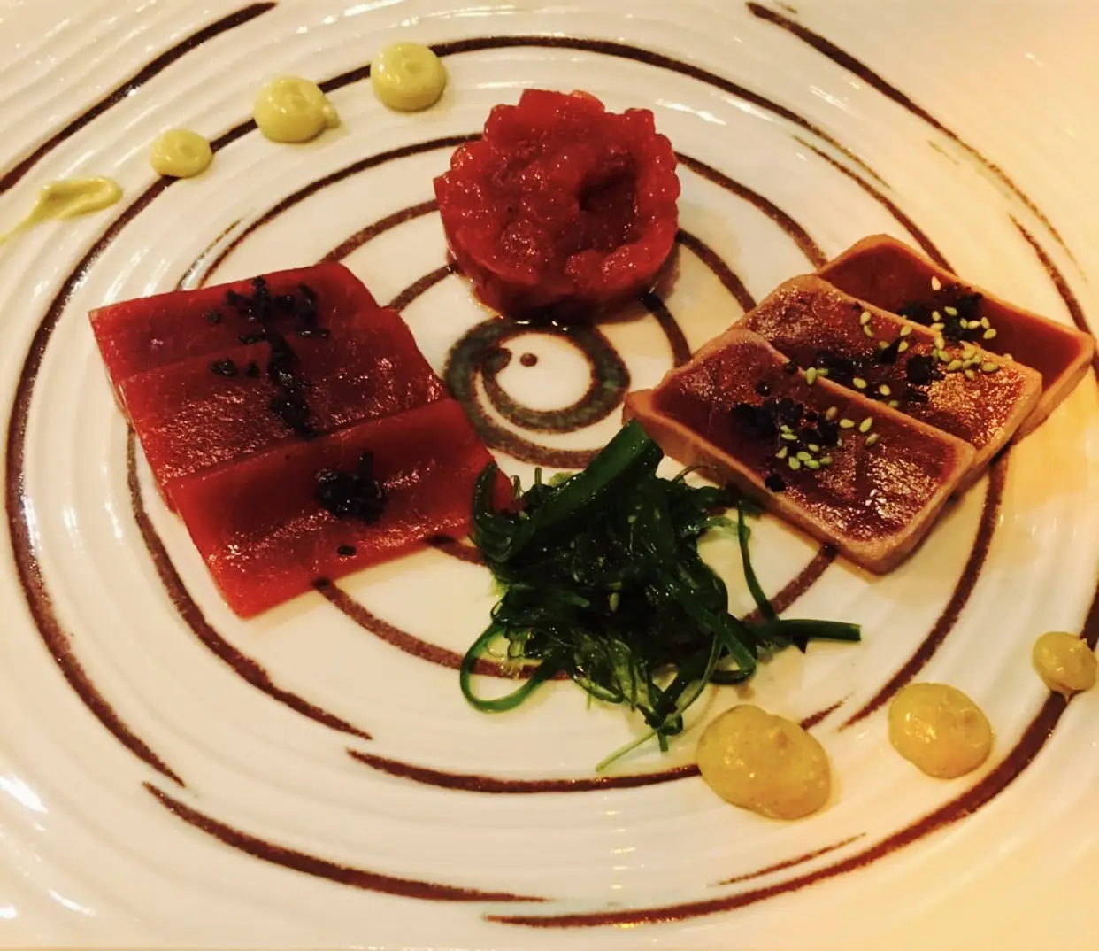 Seared tuna cubes arranged on a plate with a swirl of dark sauce and a small tartare mound.