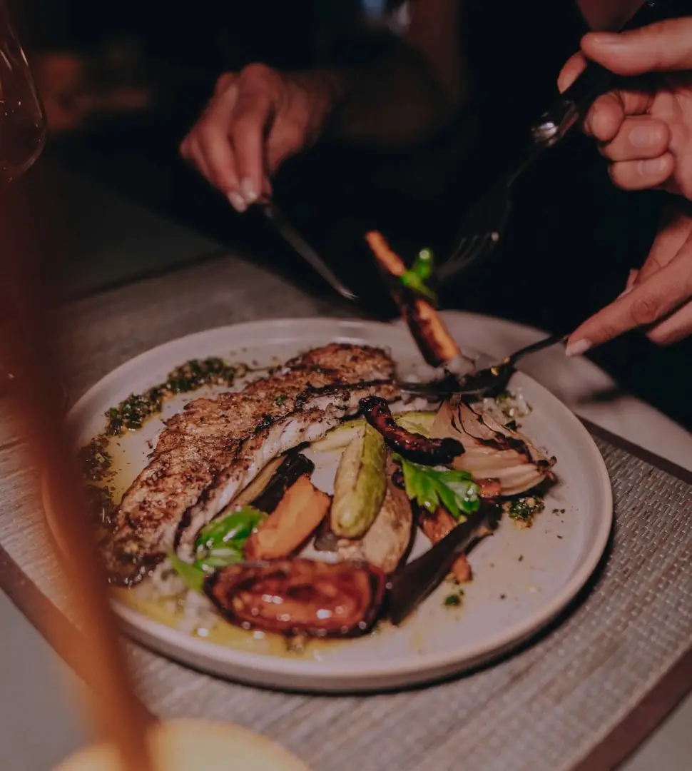 Grilled meat with charred vegetables and green herb sauce on a plate, diner cutting with fork and knife