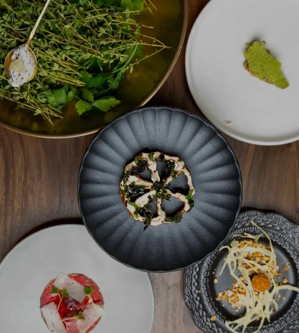 Overhead view of several small plated tasting dishes on a wooden table, including tartare and herb garnishes