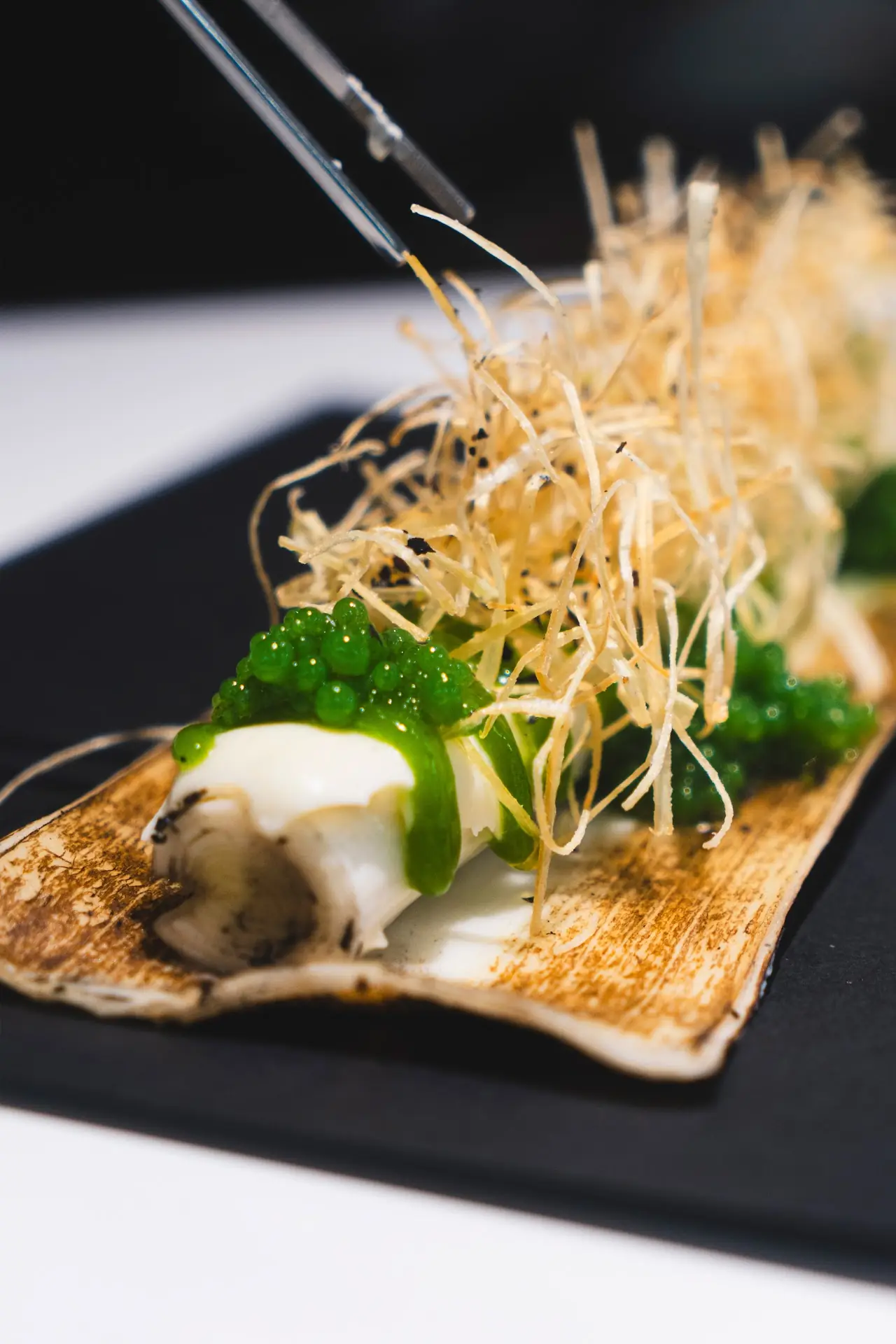 A seared scallop dish garnished with fried leek strands, green pearls, and herbs on a dark plate.