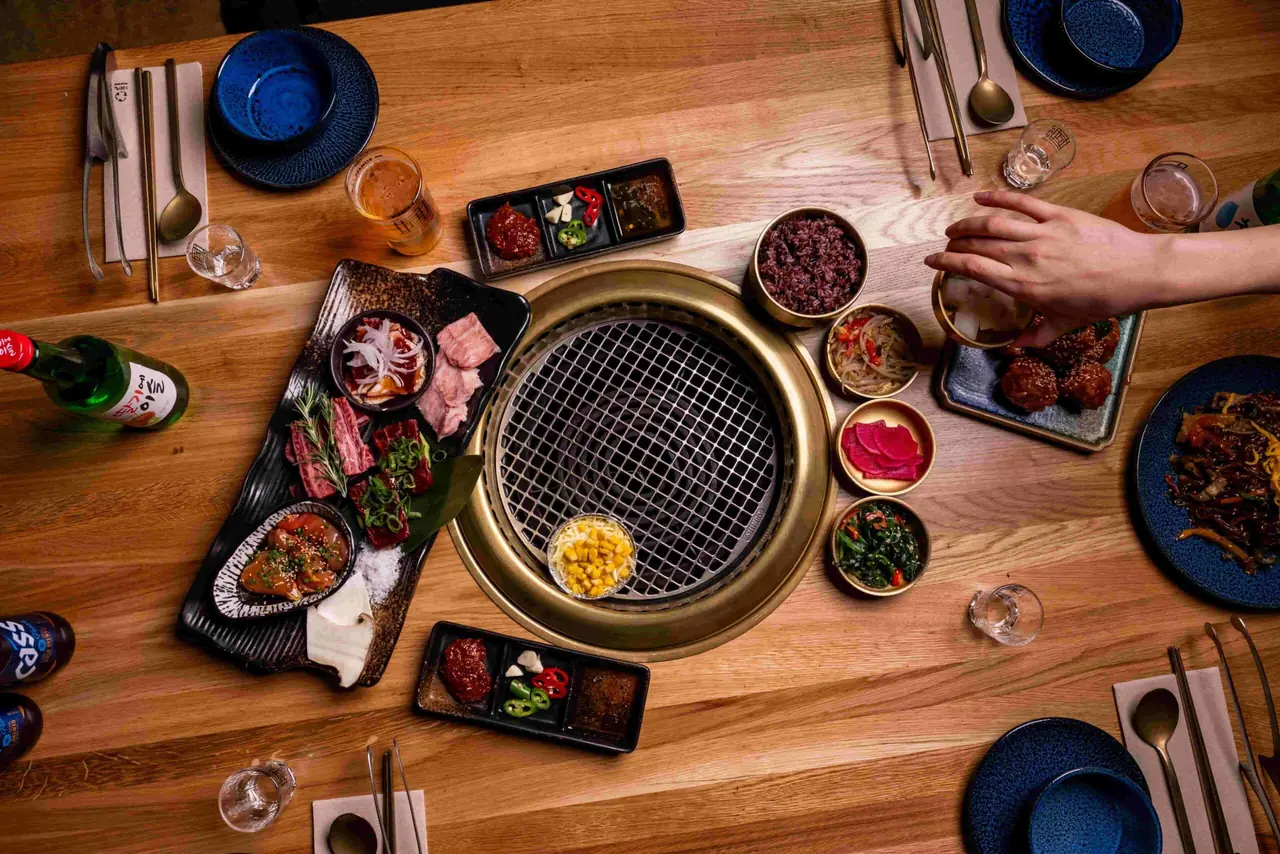 Overhead view of a Korean BBQ tabletop grill spread with marinated meats, purple rice, banchan bowls, corn, and fried chicken, GYOJASANG Korean BBQ & Bar
