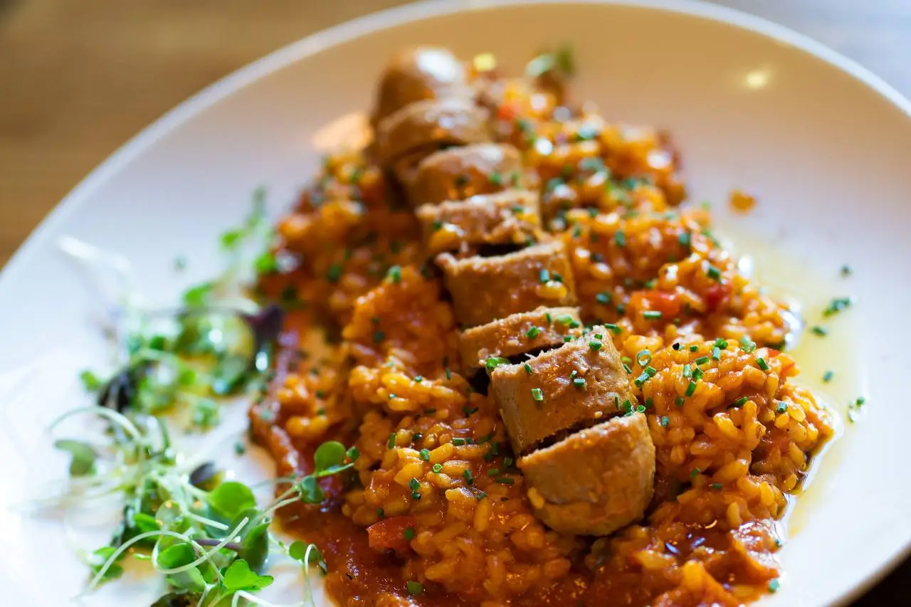 Tomato-red rice topped with sliced vegan sausage, chives and microgreens on a white plate