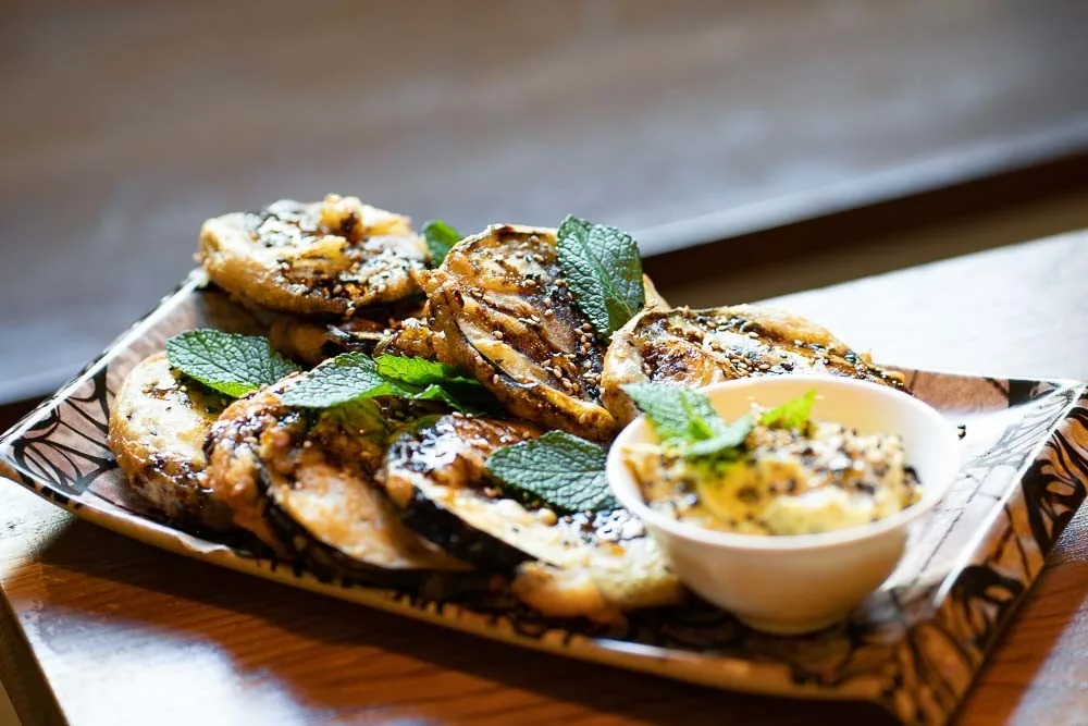 Grilled aubergine slices with fresh herbs and a small bowl of dip on a wooden board
