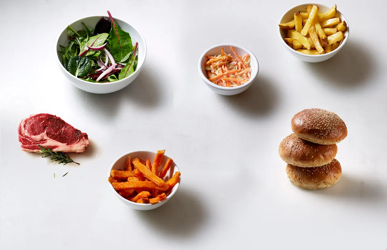 Raw seasoned steak, sesame seed burger buns stacked, and small bowls of mixed greens, coleslaw, regular fries, and sweet potato fries on a white surface, Hamburguesa Nostra