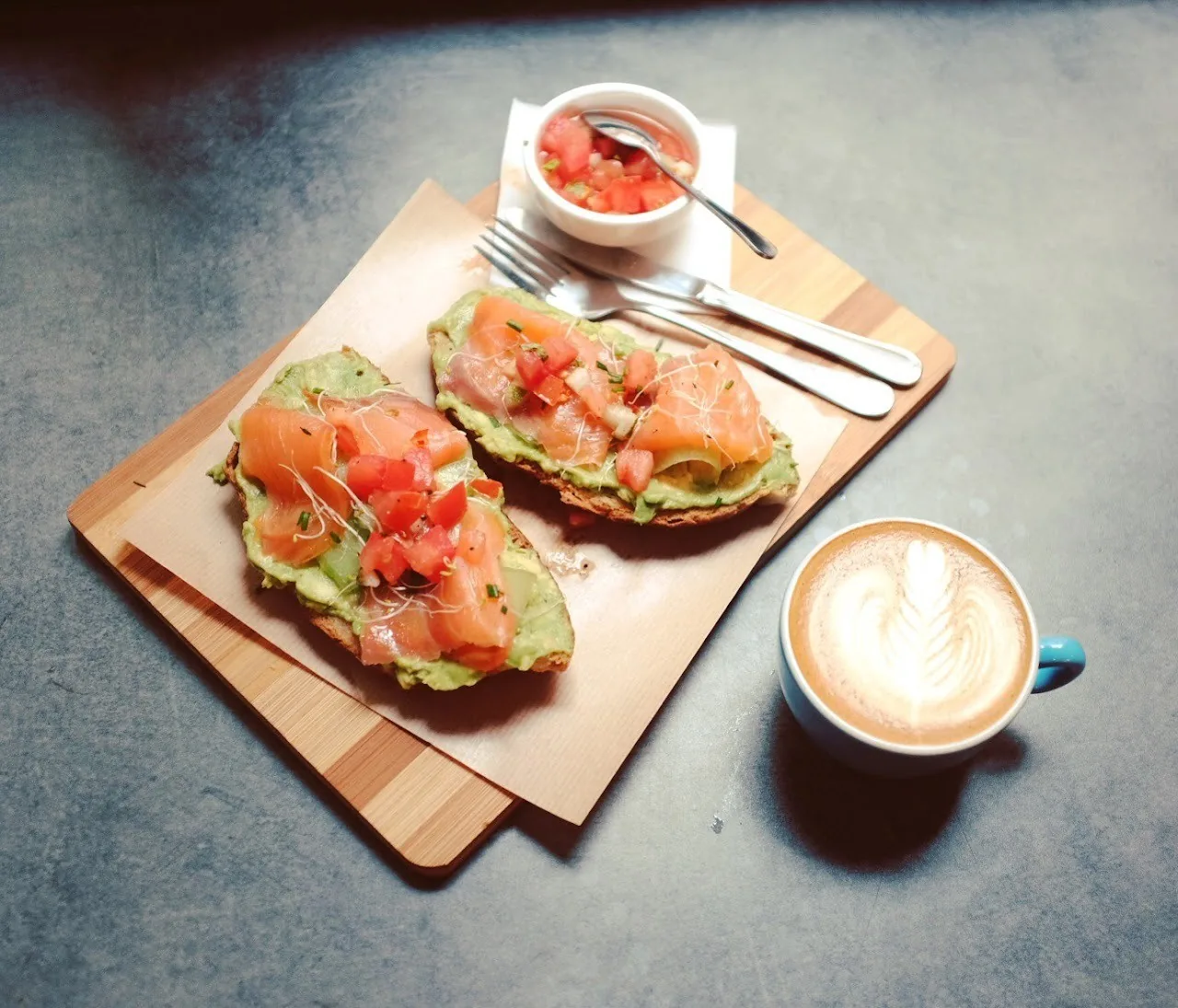 Two open toasts with avocado and salmon on a wooden board next to a latte in a blue cup