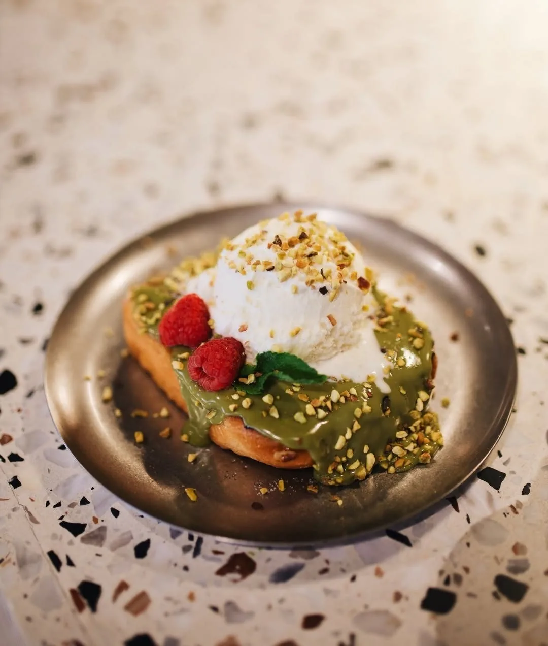A slice of toast topped with cream, berries and pistachio on a metal plate
