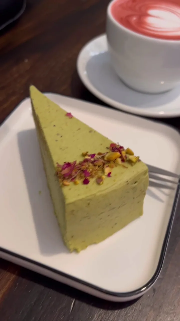 A slice of pistachio cake with rose petals beside a latte