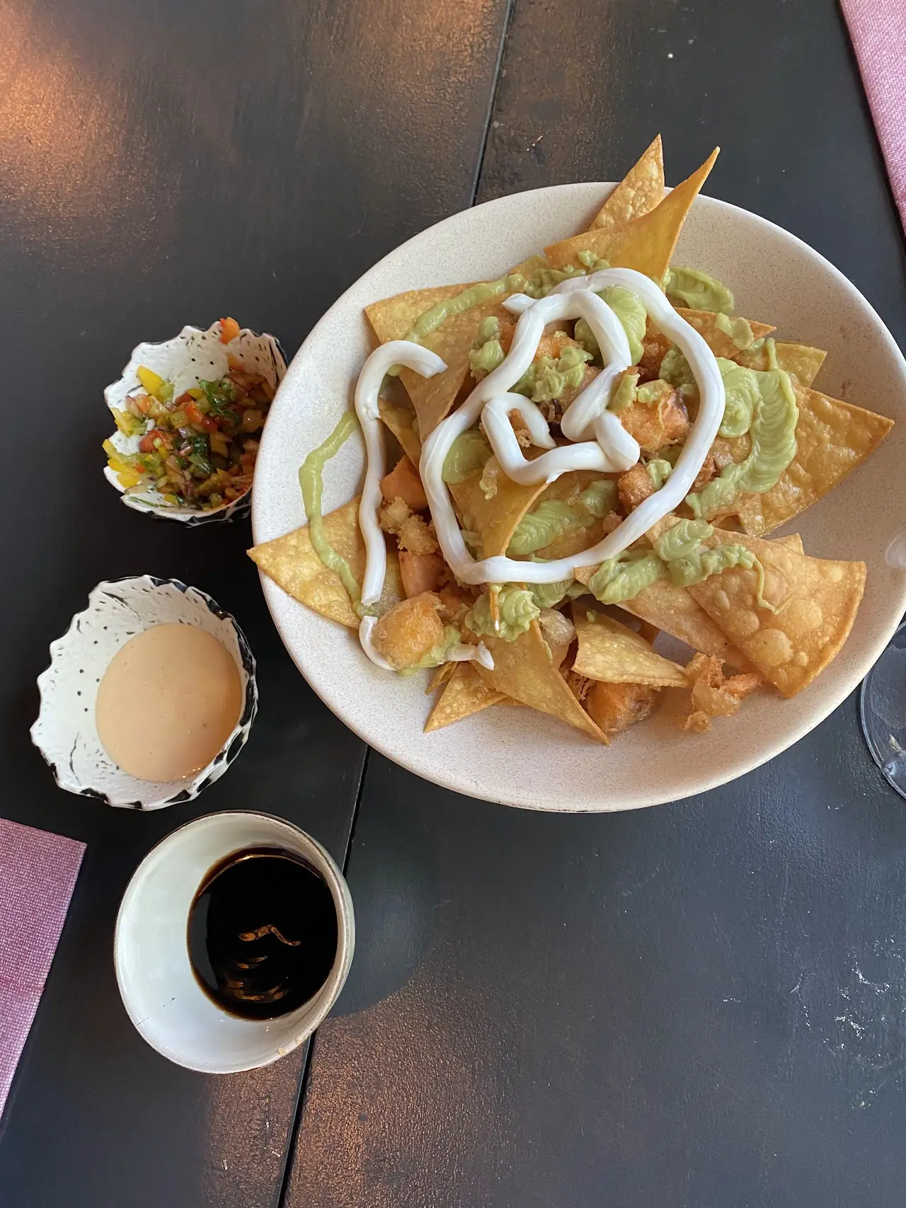 A plate of nachos topped with guacamole and a drizzled sauce, served with small side bowls.