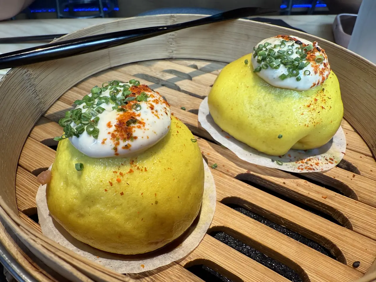 Steamed yellow bao buns topped with creamy sauce, chives and chili in a bamboo steamer