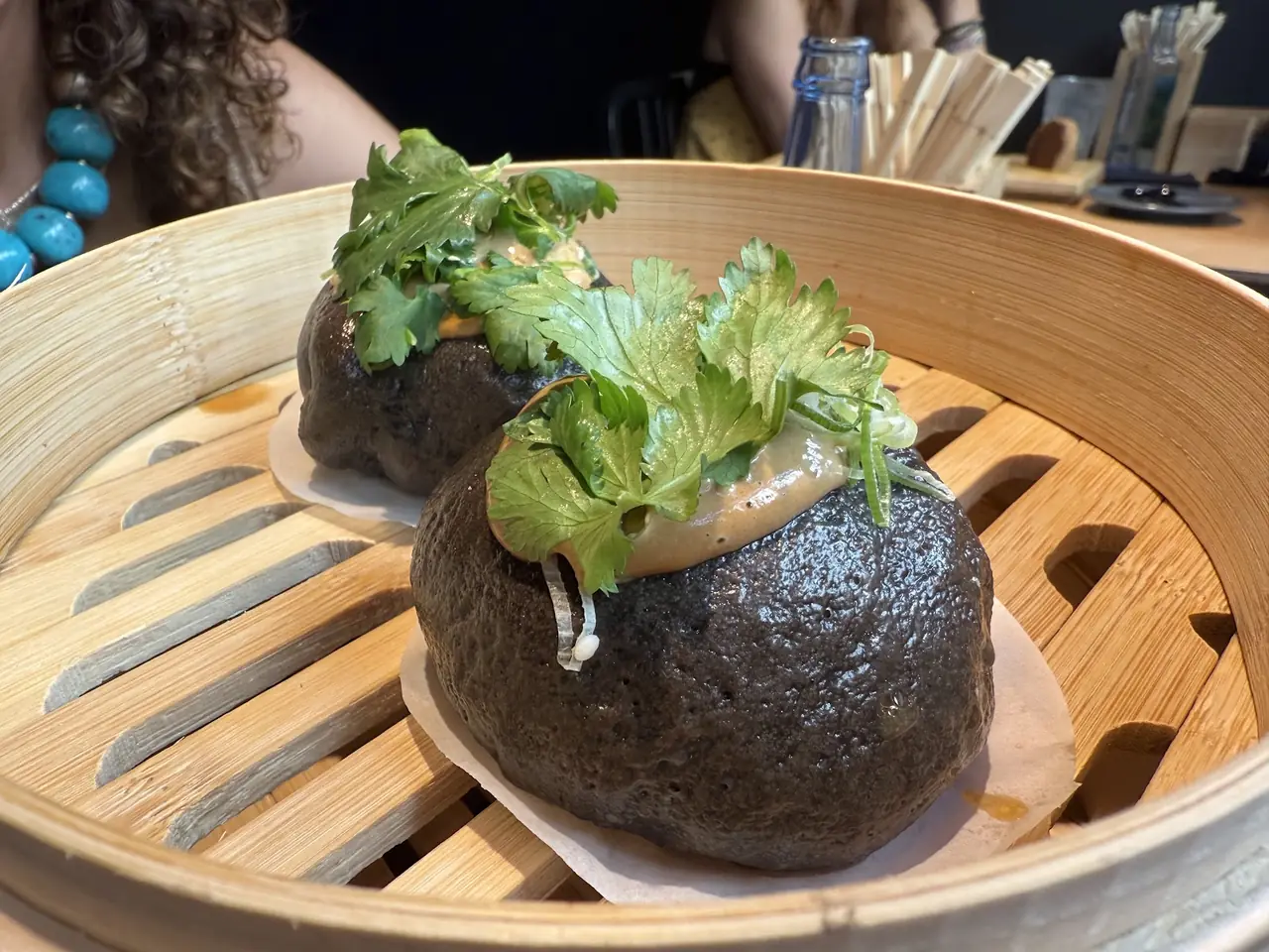 Black bao buns topped with sauce and cilantro in a bamboo steamer