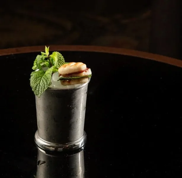 A pewter julep cup packed with crushed ice, a large mint sprig and a caramelised biscuit garnish, on a black table in a dark bar room