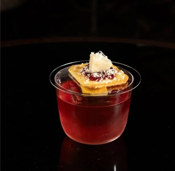 A short tumbler of clear red cocktail with a crisp wafer and a block of white cheese balanced on the rim, on a black table