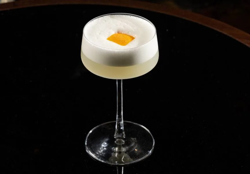 A coupe glass of pale cocktail under a thick white foam head with a small square of orange peel set into it, reflected in a black table