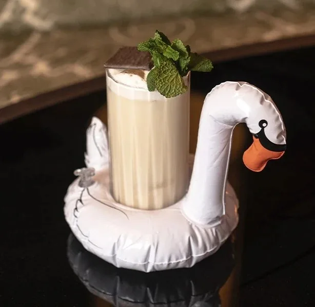 A tall glass of pale, creamy cocktail topped with a square of dark chocolate and a mint sprig, sitting in an inflatable swan drinks float on a black table