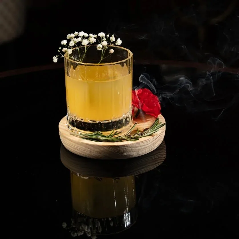 A cocktail in a tumbler with small white flowers on a wooden coaster with red flowers beside it