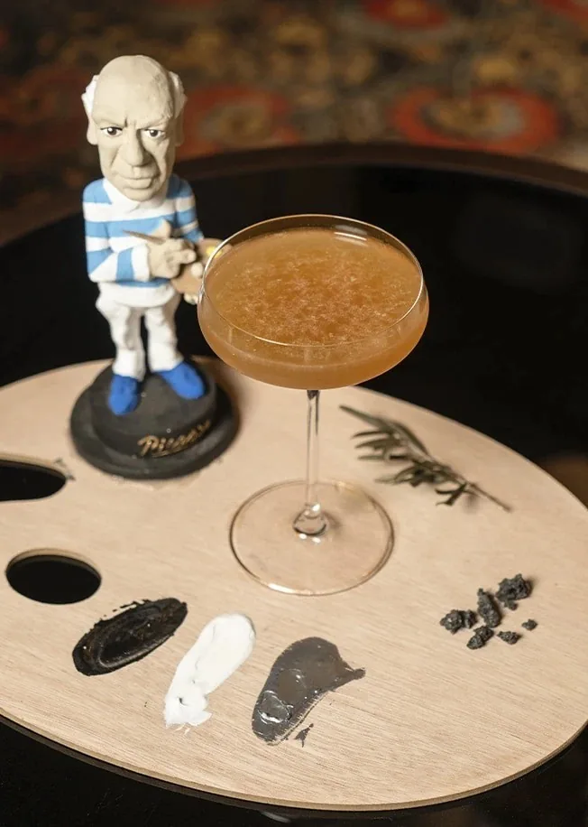 A coupe cocktail on a wooden palette beside a small figurine