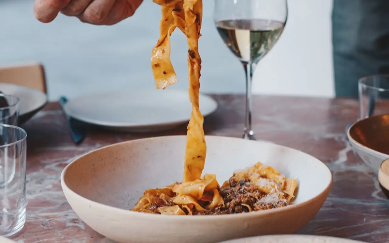 Pappardelle in ragu being lifted from a stoneware bowl over a marble table with a glass of white wine alongside