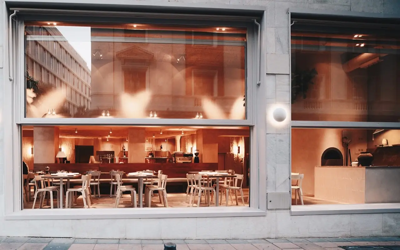 Restaurant seen through large street windows at dusk, warm-lit dining room with pale wooden chairs, a bar and a wood-fired oven