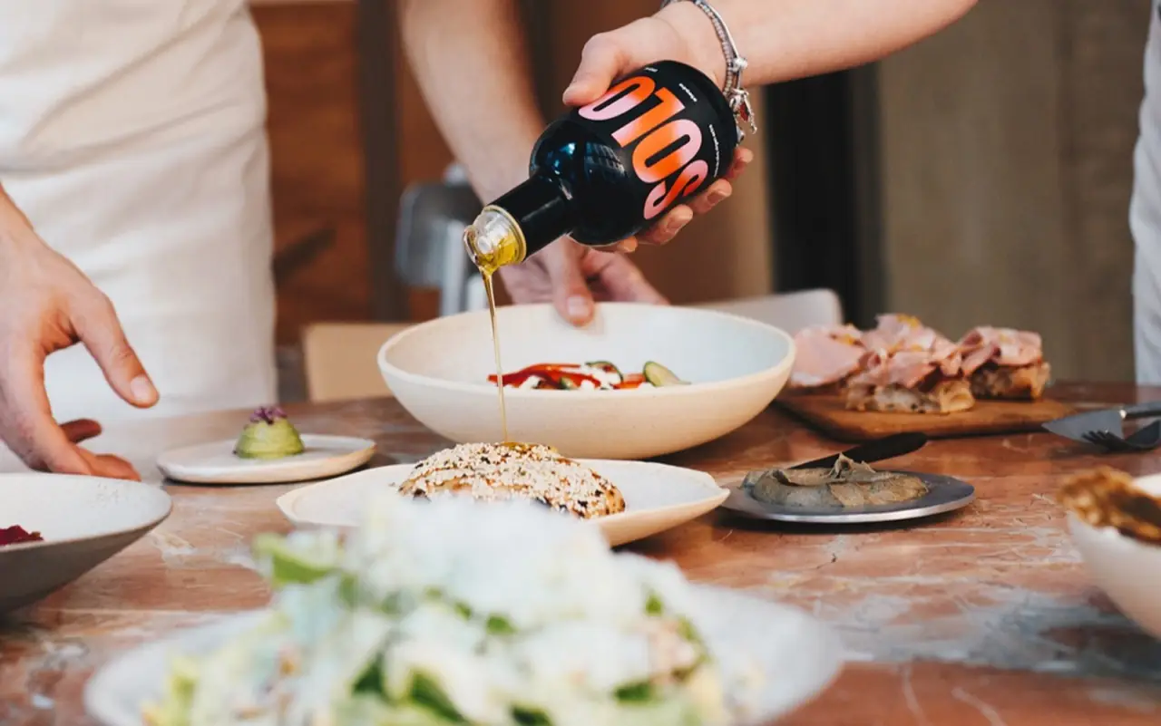 Table spread of dishes with a bottle of SOLO olive oil being poured over a salad bowl, sliced ham and a seeded flatbread