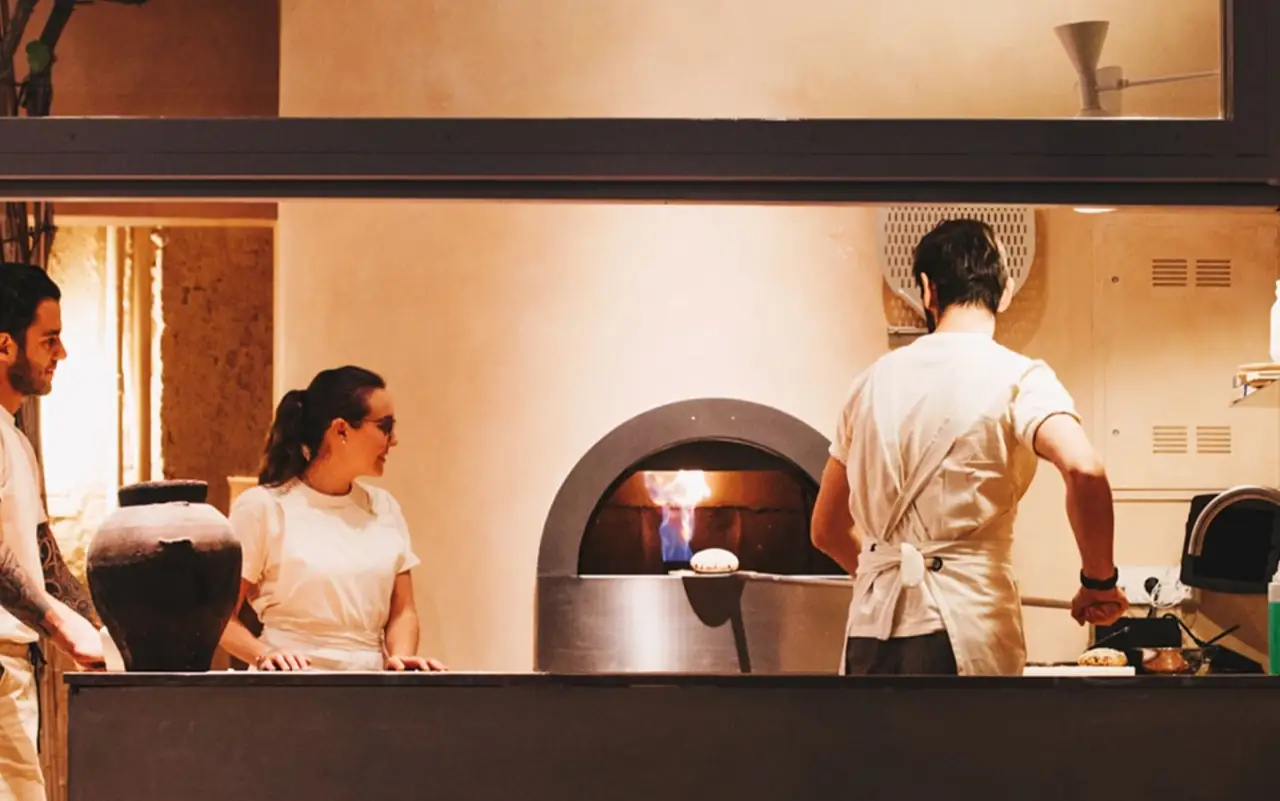 Open kitchen with a domed wood-fired oven aflame, cooks in white aprons working a flatbread and a large ceramic urn on the counter