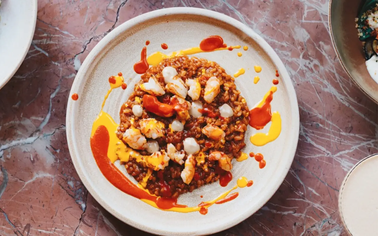 Soupy rice with prawns dressed in red and yellow sauce swirls on a stoneware plate atop a pink marble table