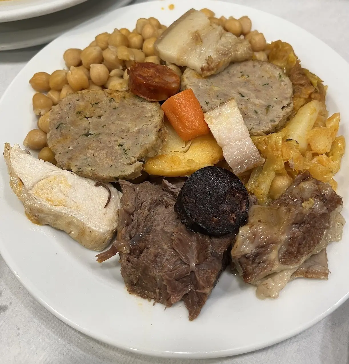 A white plate of cocido: chickpeas, cabbage, carrot, chorizo, morcilla and boiled meats