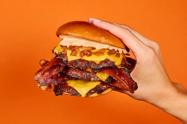 Stacked smash burger with melted cheese and crispy bacon strips held in a hand at Hideout Burger