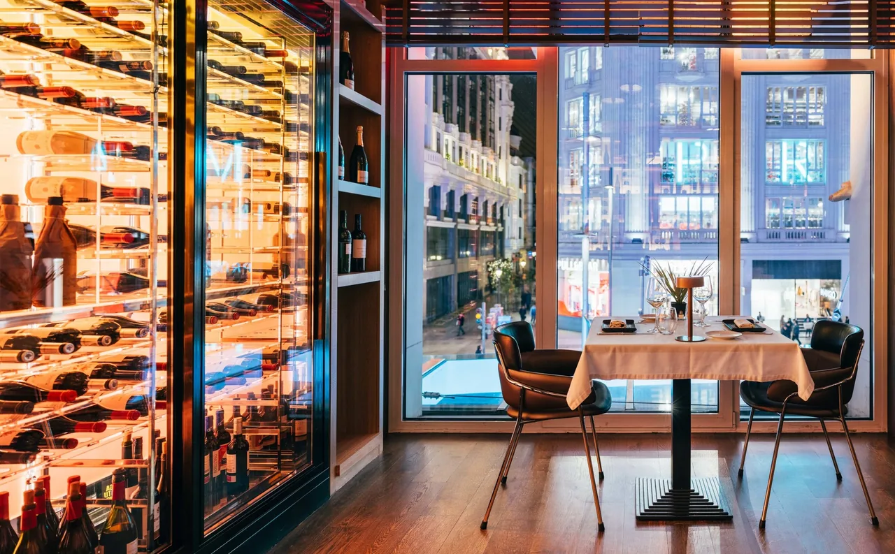 A table set for two beside tall windows overlooking a lit city street at night, with a glass wine cabinet at left