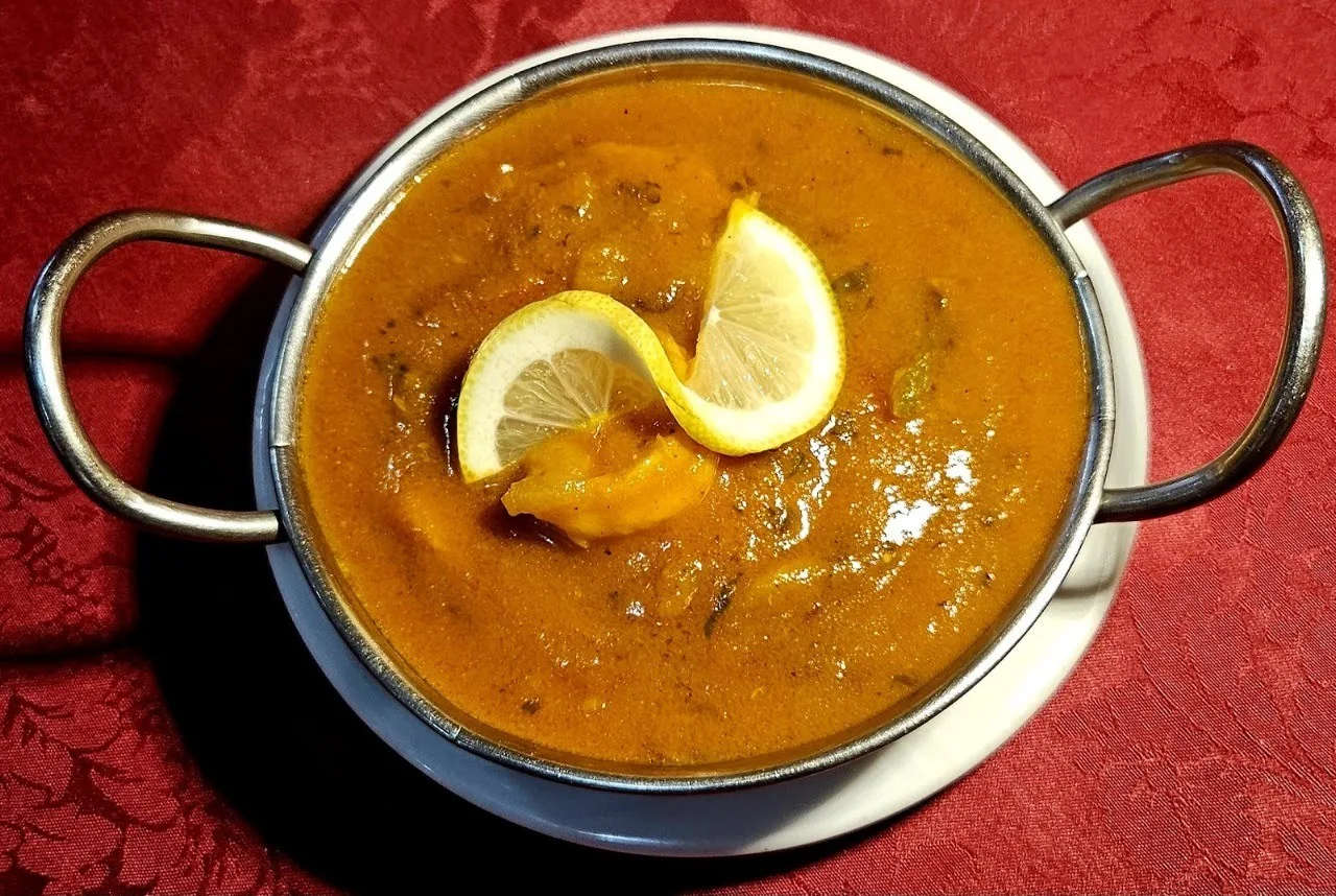 A metal handled bowl of orange curry with a twist of lemon on top, on a red tablecloth