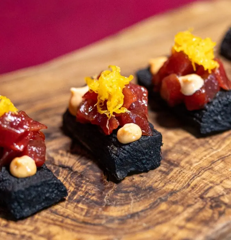 Squares of black yuca topped with spicy red tuna tartare, grated yolk and dots of sauce, on a wooden board