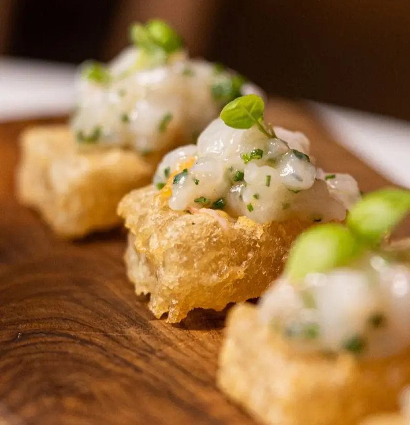Crisp tempura tapioca squares topped with langoustine tartare and spring onion, on a wooden board
