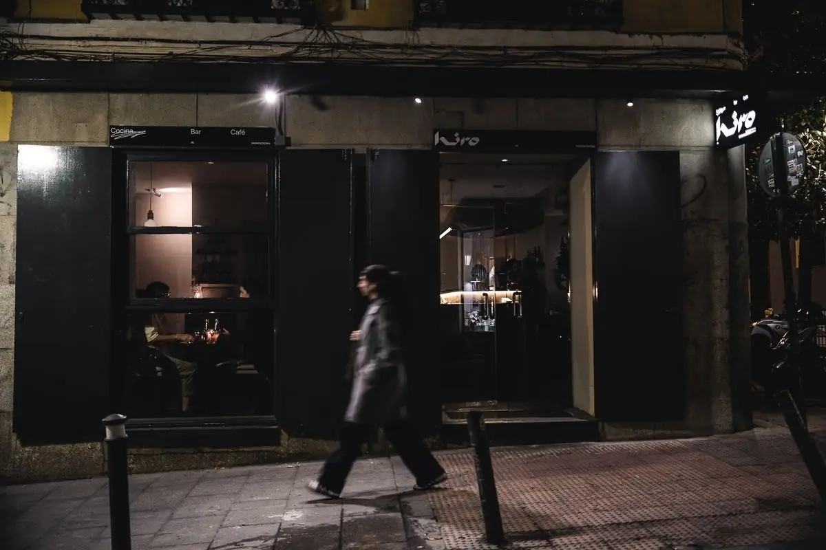 Night-time street exterior of Kiro, a black storefront signed Cocina Bar Cafe, with a passerby walking by and warm-lit interior visible through the windows