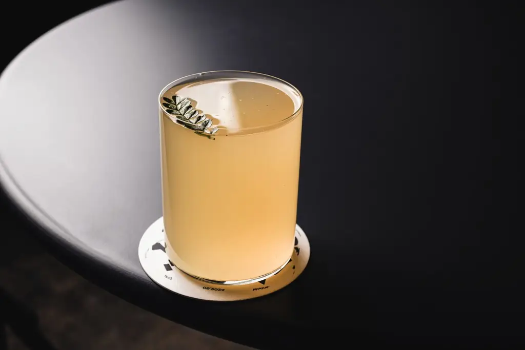 Pale cloudy cocktail in a rocks glass with a small leaf garnish, resting on a coaster on a dark round table