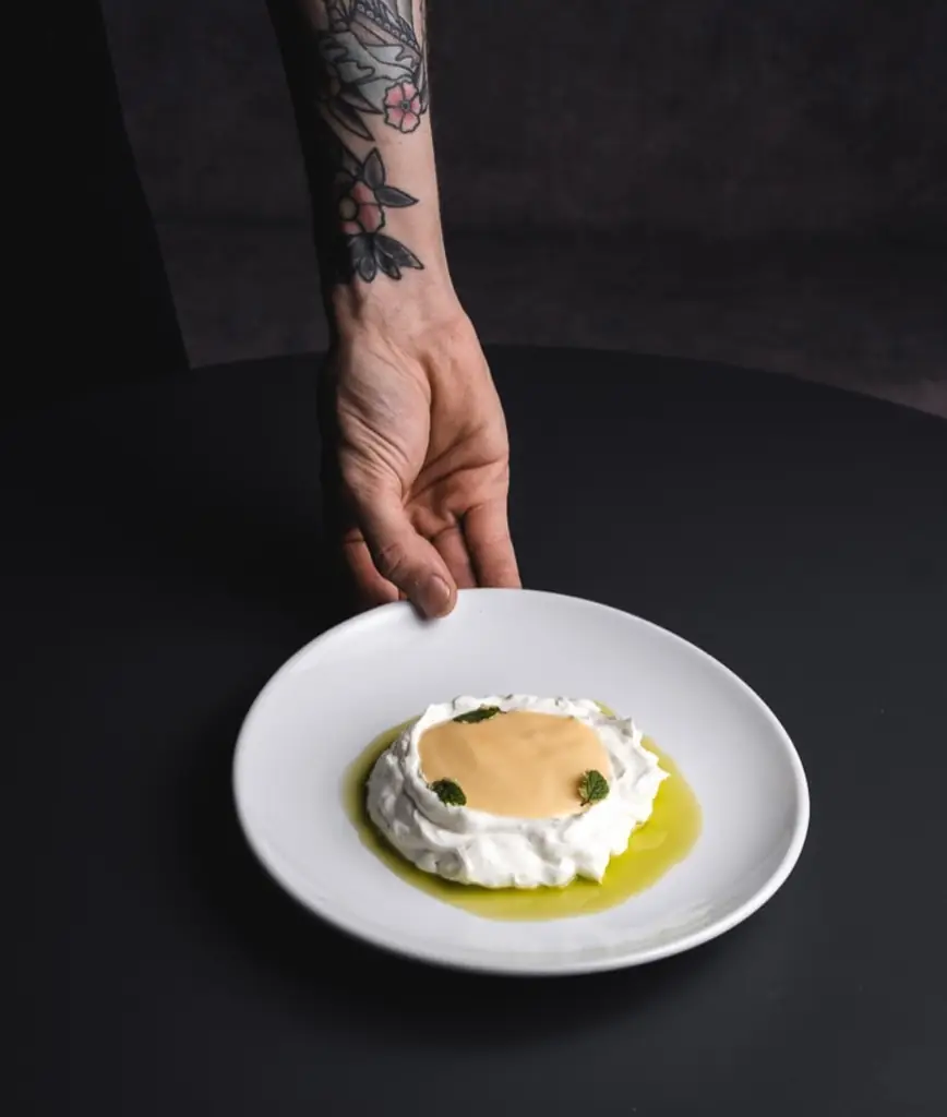 White plate of whipped cream or labneh topped with a smooth sauce, mint leaves and olive oil, being set down by a tattooed hand
