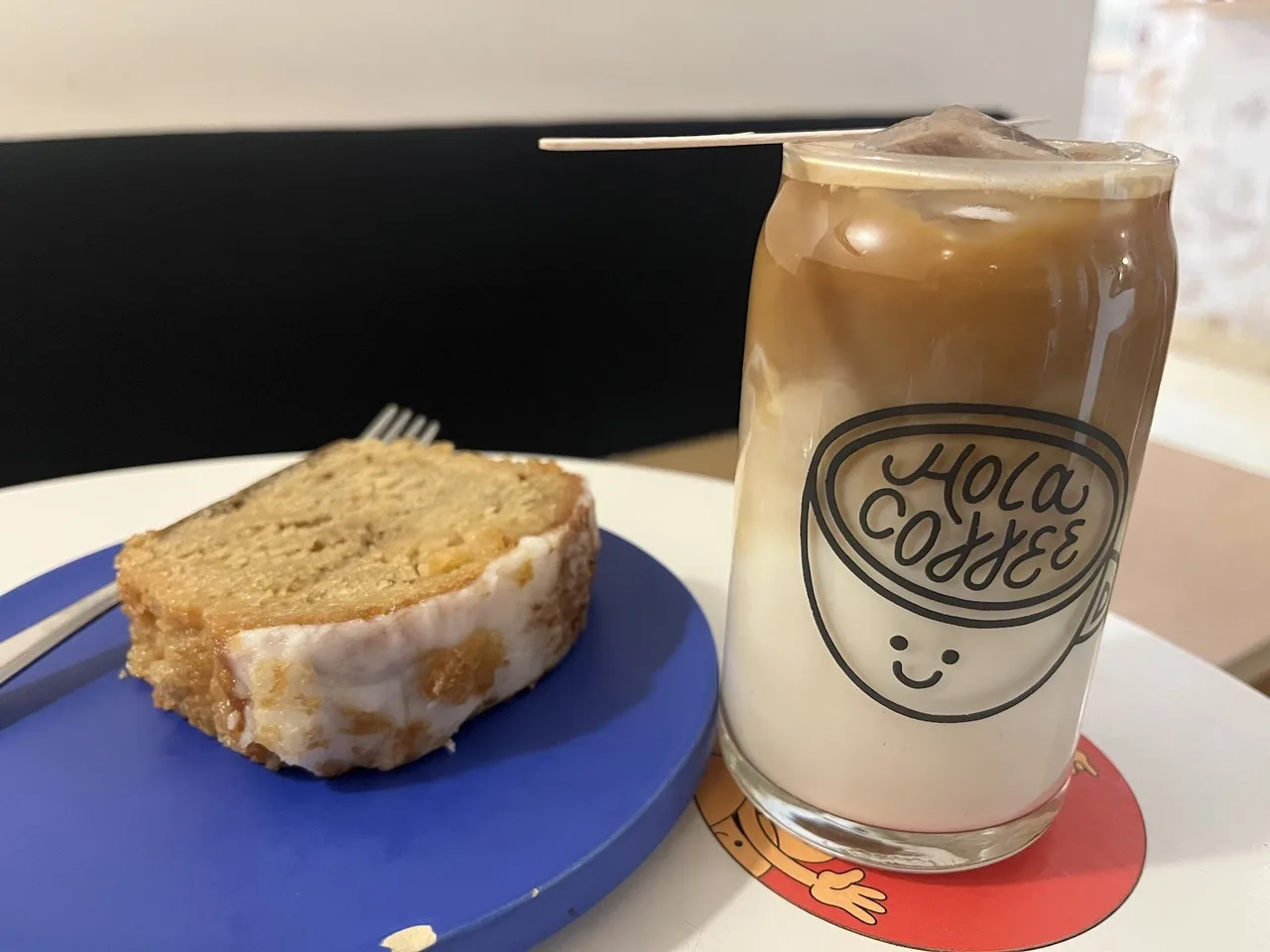 An iced coffee in a glass with a smiling cup logo beside a slice of loaf cake on a blue plate