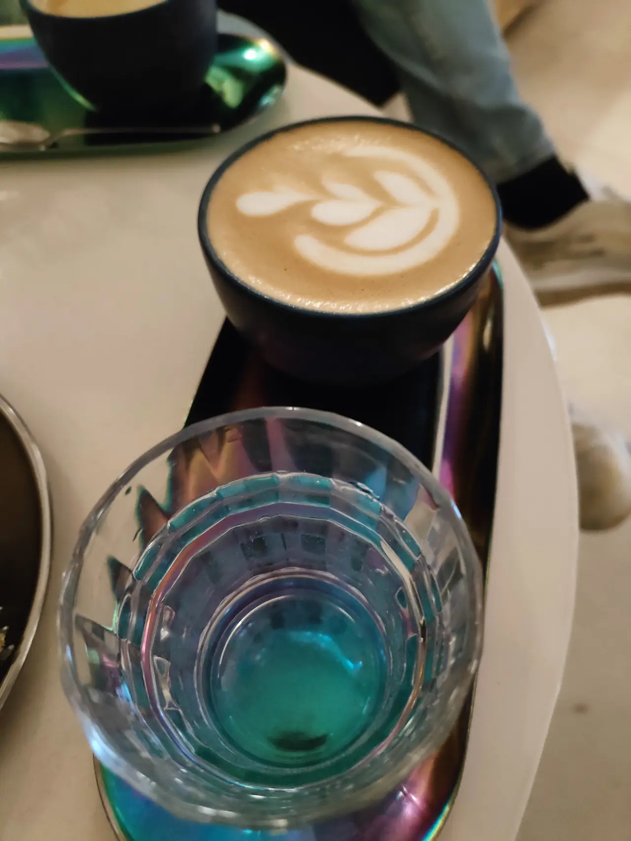 Latte with leaf latte art in a black cup next to a water glass on a tray