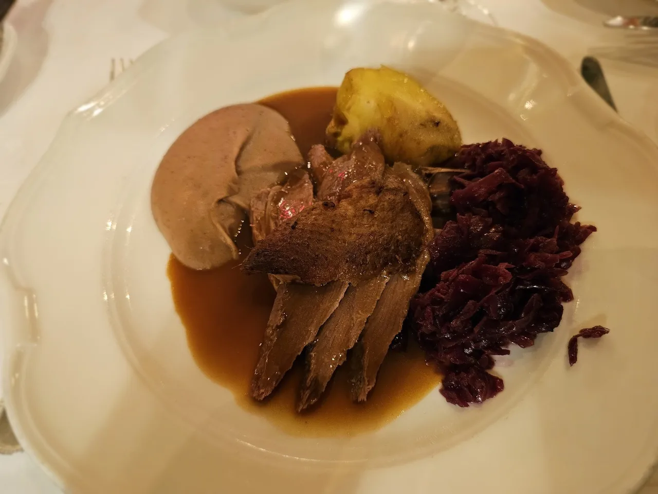 Slices of dark roast meat with a smooth pale portion, a lemon wedge and red cabbage on a plate with sauce