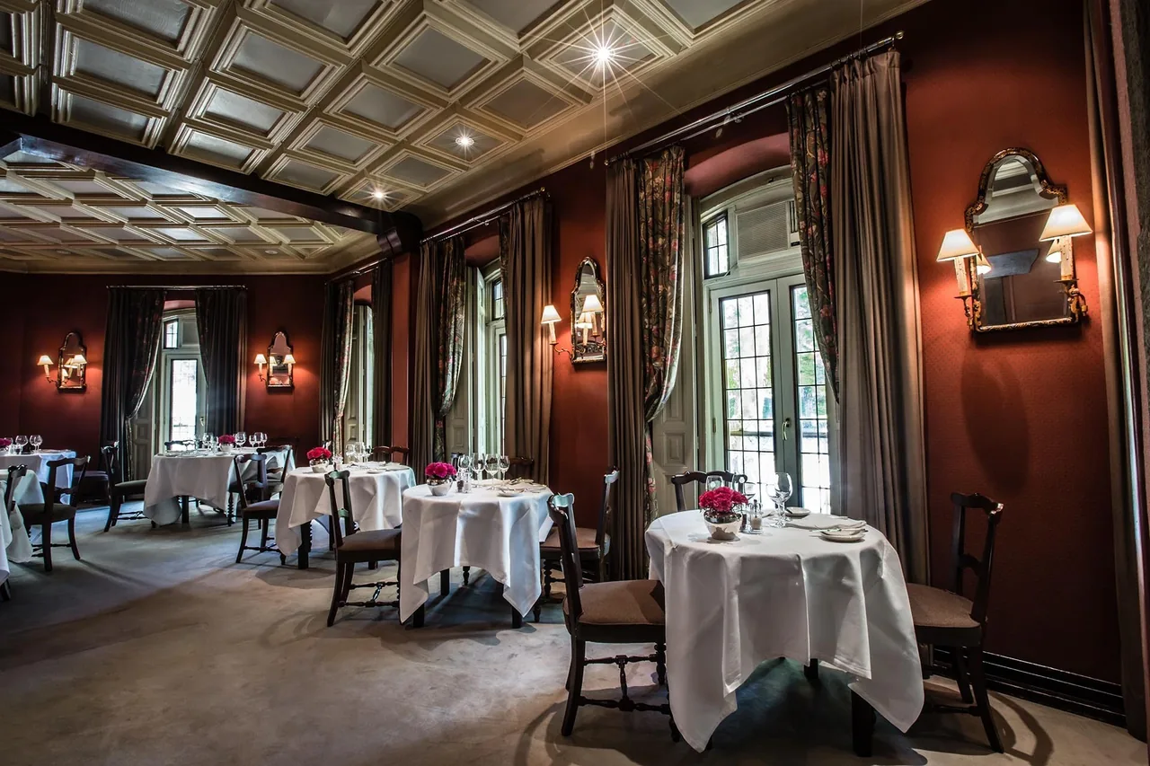 A dining room with red walls, coffered ceiling, tall windows and round tables set with white cloths.