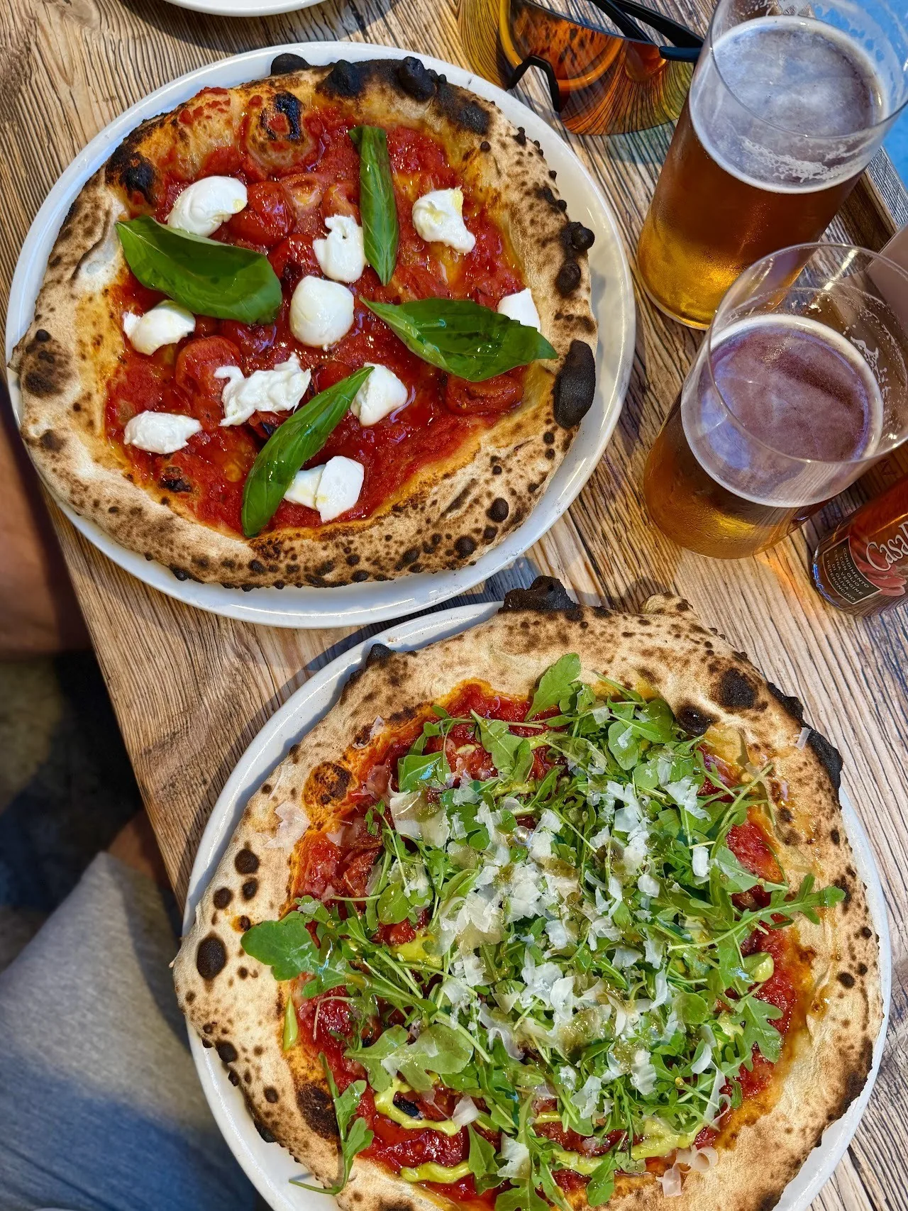 Two pizzas seen from above on a wooden table, one with white cheese and basil and one with rocket, next to glasses of beer