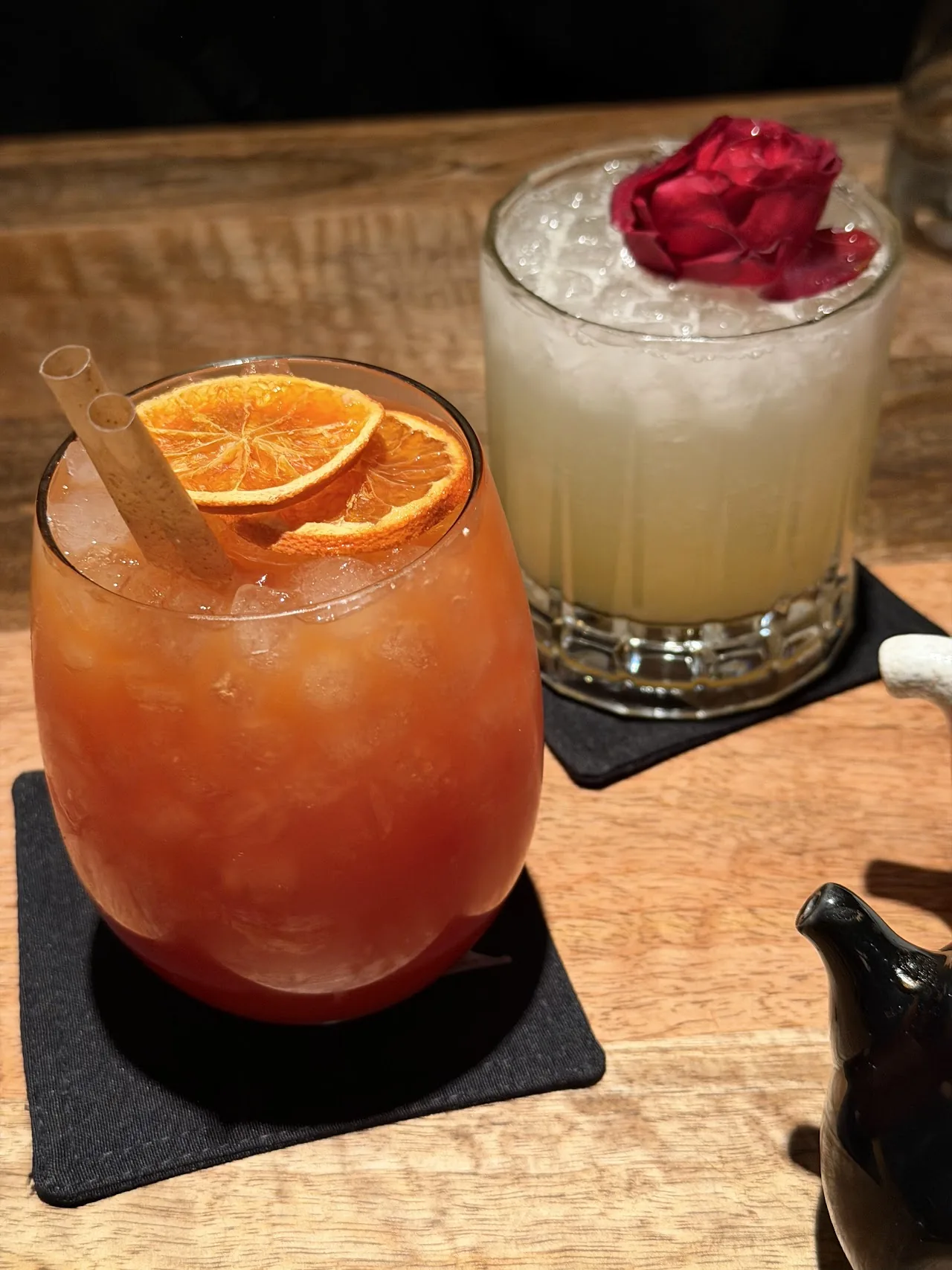 Two cocktails, one orange with an orange wheel garnish and one pale with a rose petal