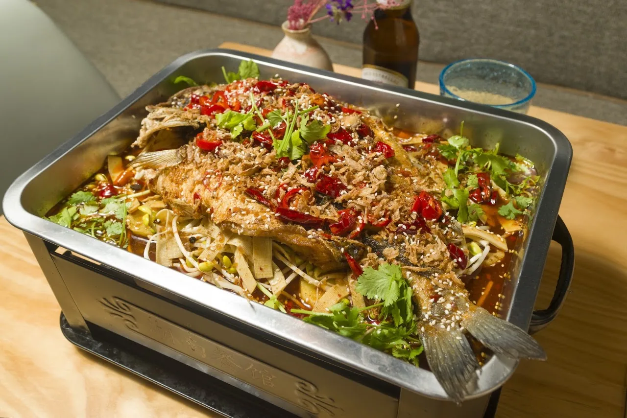 A whole fish in savory sauce with greens in a metal tray